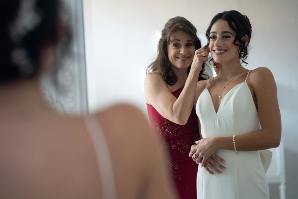 A mother helps her daughter get ready for her wedding smiling as she adjusts her earrings.jpg