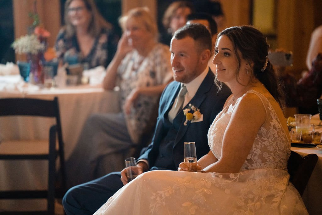 A newly married couple takes in the speeches at their wedding reception They are beaming and look so happy.jpg