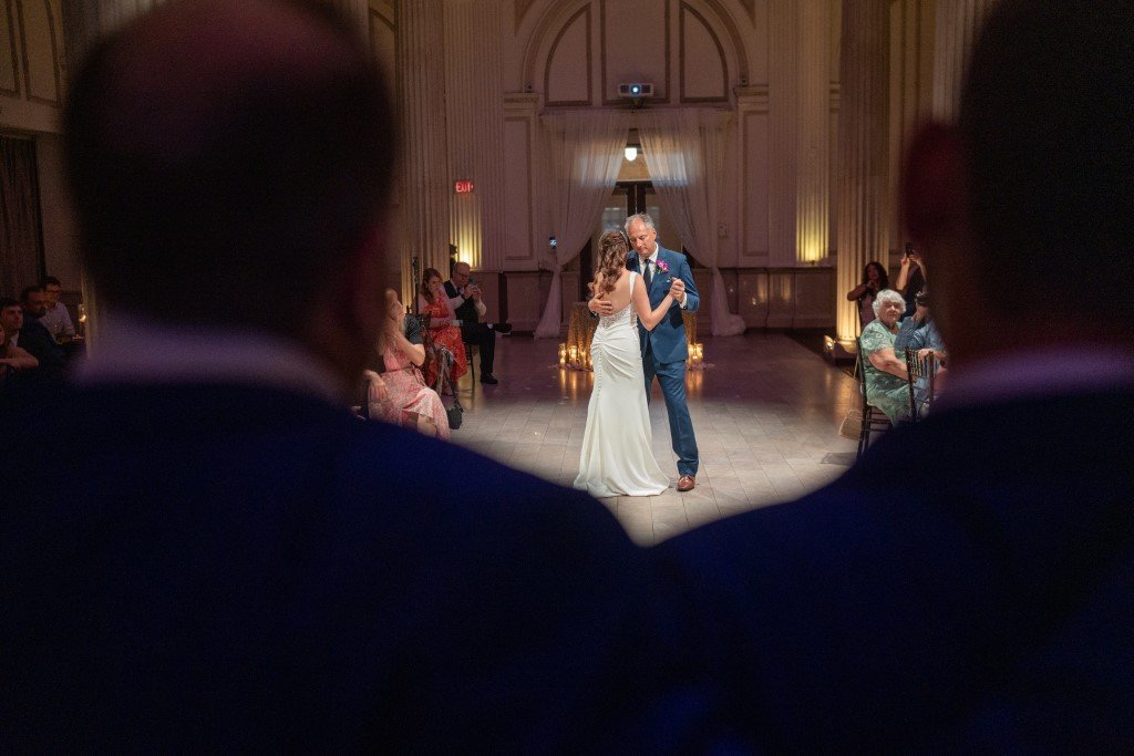 A newlywed couple shares their first dance as husband and wife surrounded by family and friends in a beautifully lit hall.jpg