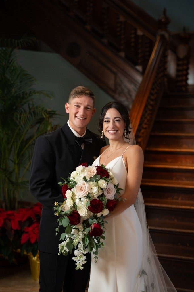 A newlywed couple smiles in front of a staircase holding a bouquet of white and red flowers.jpg