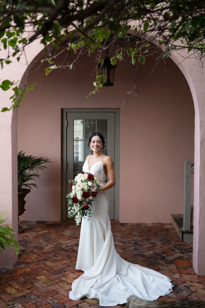 Bride in a white dress holds a bouquet standing under an archway adorned with greenery.jpg