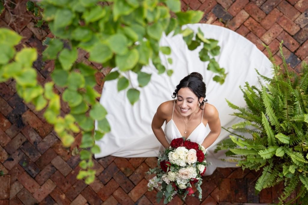 Bride smiling radiantly with a bouquet in her hands seen from above on a brick path surrounded by greenery.jpg