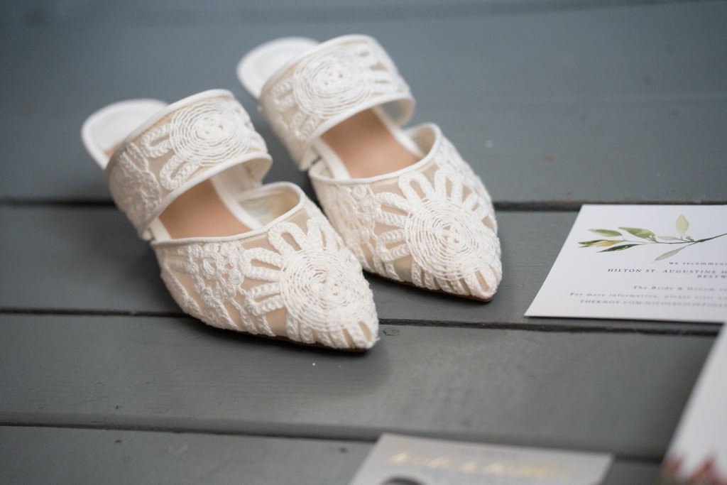 Elegant white lace bridal shoes placed on a wooden floor next to wedding invitations.jpg