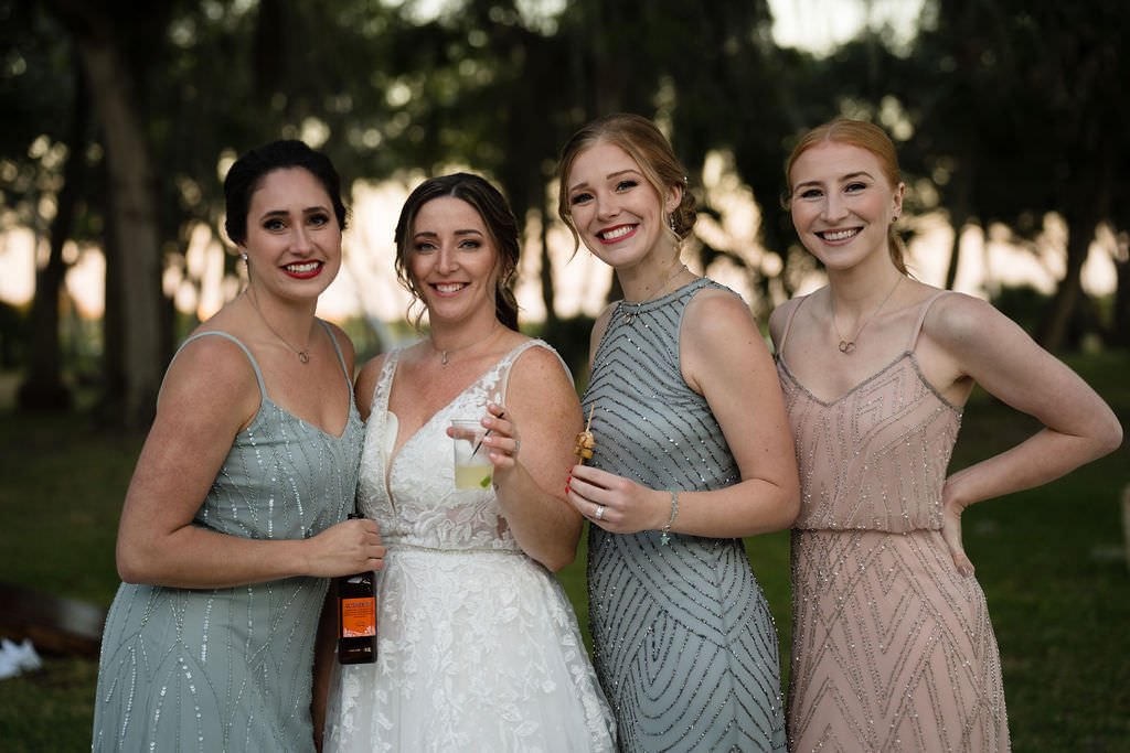 Four women in formal dresses smiling and posing outdoors with one in a wedding dress holding a drink.jpg
