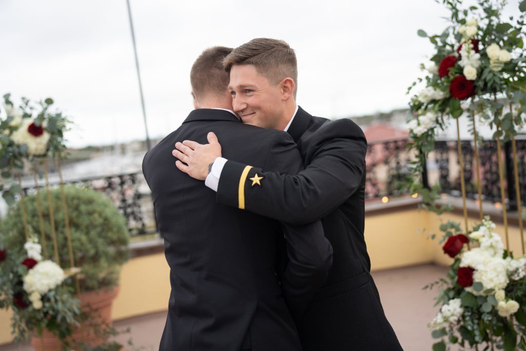 Two men share a heartfelt hug at an outdoor event surrounded by floral arrangements.jpg